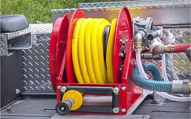 A yellow hose coiled on a red reel, mounted on a metallic surface with attached pipes and a nozzle, ready for use ASAP.